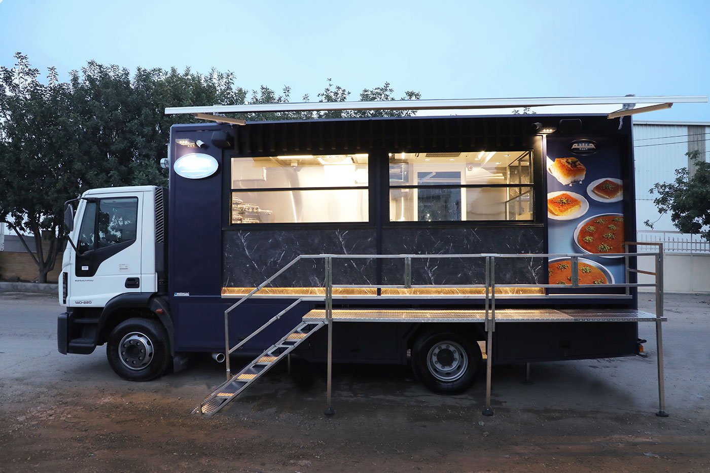 The finished Habibah food truck at dusk: a navy box body on a white-cab truck, with lit serving windows, a fold-down awning, and a stainless steel serving deck with stairs.