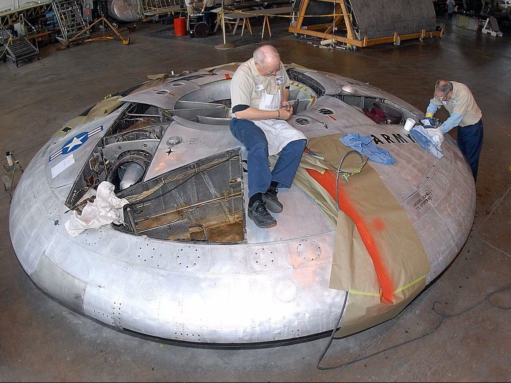 Two technicians working on a preserved Avrocar inside a museum restoration hangar.